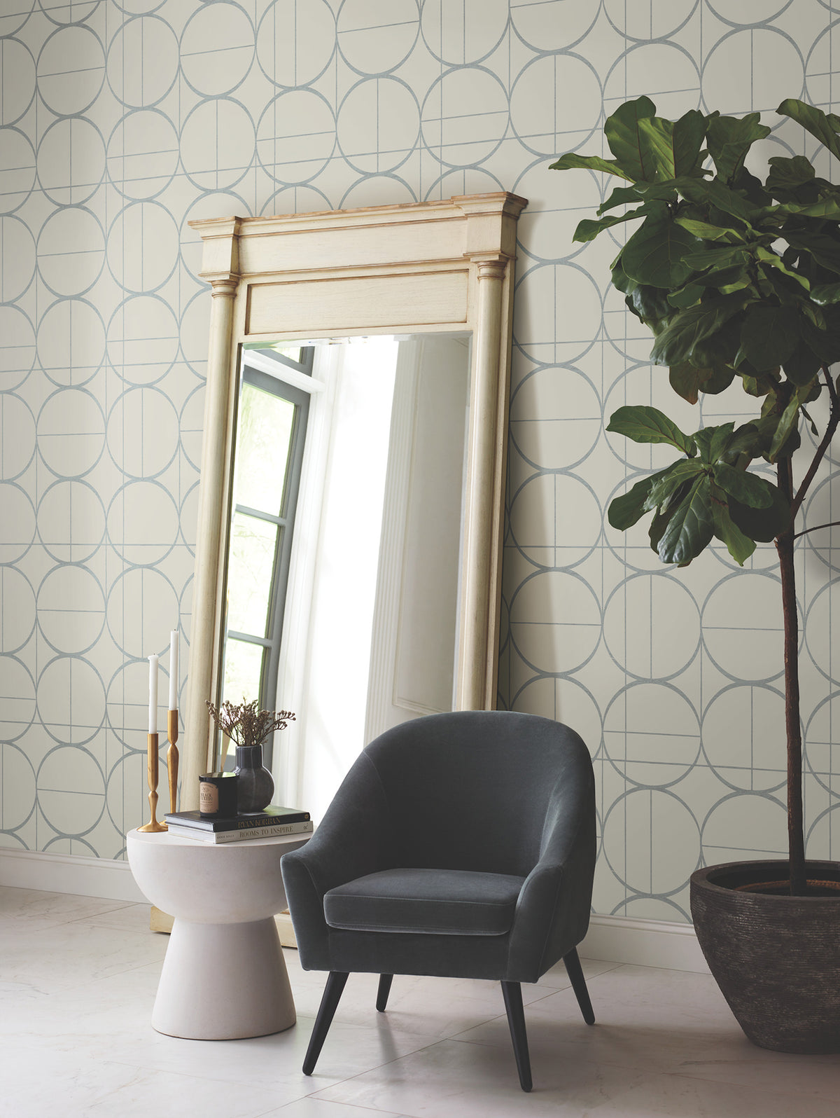 A large mirror with a cream frame stands against a wall covered in York Wallcoverings’ Sun Circles Wallpaper. A dark armchair and small round side table with decor sit nearby, next to a tall potted plant. Light streams in from a window.