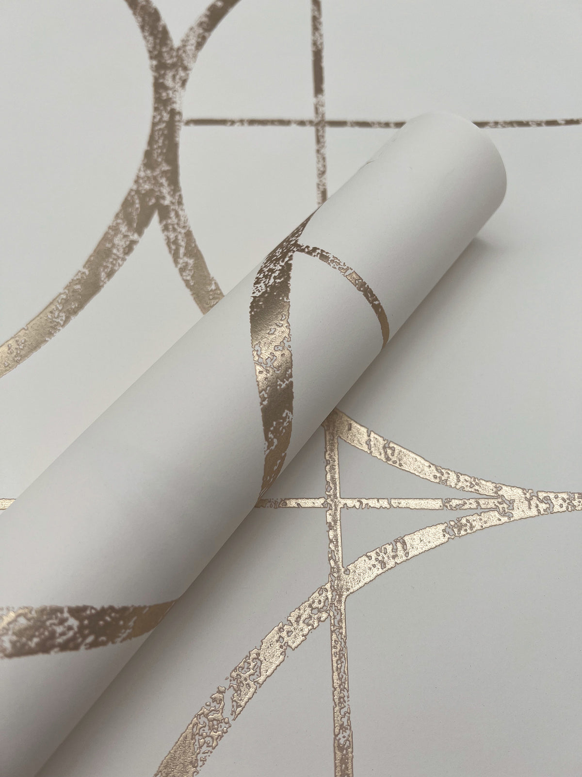 A roll of Sun Circles Wallpaper by York Wallcoverings, featuring shiny gold geometric lines on a white background, is partially unrolled on a matching patterned surface.