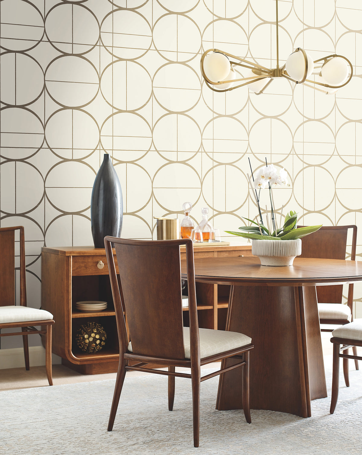 Mid-century modern dining room with a wooden table and chairs, a sideboard featuring a tall vase and gold decor, York Wallcoverings’ Sun Circles Wallpaper as an accent, a white orchid centerpiece, and a brass chandelier overhead.