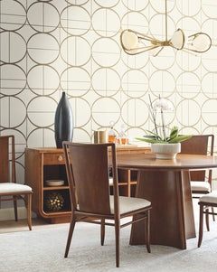 Mid-century modern dining room with a wooden table and chairs, a sideboard featuring a tall vase and gold decor, York Wallcoverings’ Sun Circles Wallpaper as an accent, a white orchid centerpiece, and a brass chandelier overhead.