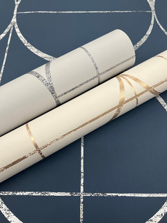 Two rolls of Sun Circles Wallpaper by York Wallcoverings—one showcasing silver geometric lines and the other gold—are positioned on a blue background adorned with a coordinating silver geometric pattern.