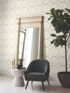 A modern room featuring the Sun Circles Wallpaper by York Wallcoverings, which displays a geometric gold circle pattern on a white background. The space includes a large mirror, a dark gray armchair, a white side table adorned with decor items, and a tall potted plant, all illuminated by natural light.
