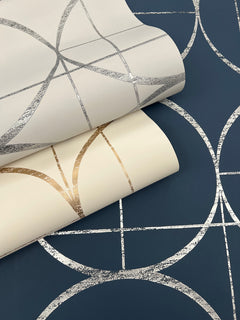 Two rolls of York Wallcoverings’ Sun Circles Wallpaper—one in cream with metallic gold geometric lines and another in cream with metallic silver lines—are displayed on a dark blue surface featuring a silver geometric circle pattern.