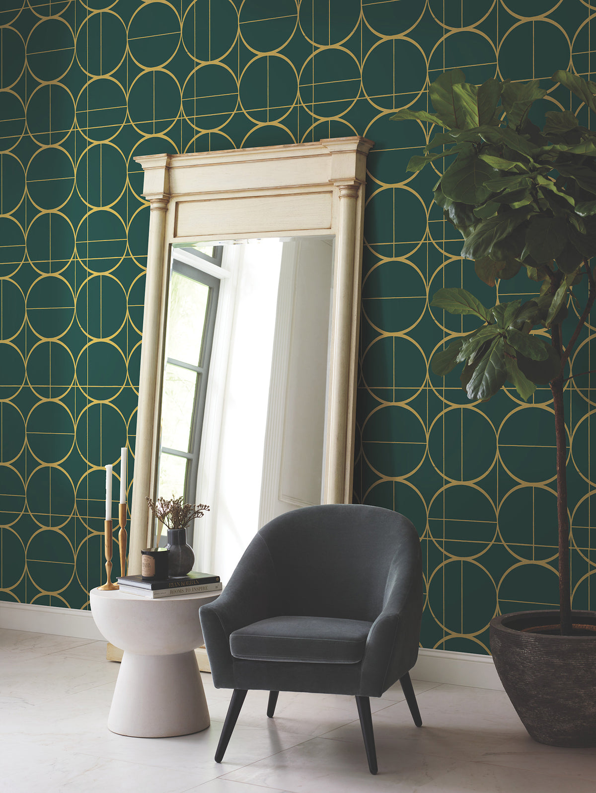 A modern room featuring York Wallcoverings’ Sun Circles Wallpaper in green with a gold geometric pattern, complemented by a large ornate mirror, a small round side table with decor, a dark gray armchair, and a potted plant.