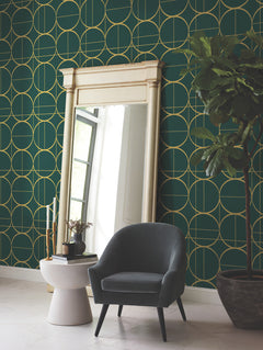 A large mirror with a cream frame stands against a green wall adorned with York Wallcoverings’ Sun Circles Wallpaper in gold geometric patterns. A gray armchair sits nearby, next to a round side table with decor, and a tall potted plant is on the right.