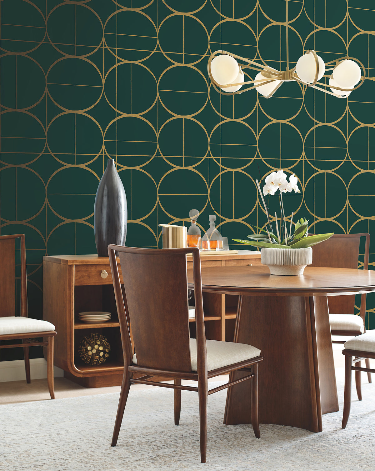 A mid-century modern dining room featuring a wooden table and matching chairs, highlighted by York Wallcoverings’ Sun Circles Wallpaper in green and gold geometric patterns. The space is completed with a white orchid centerpiece, a sideboard adorned with vases, and a modern chandelier showcasing round white lights.