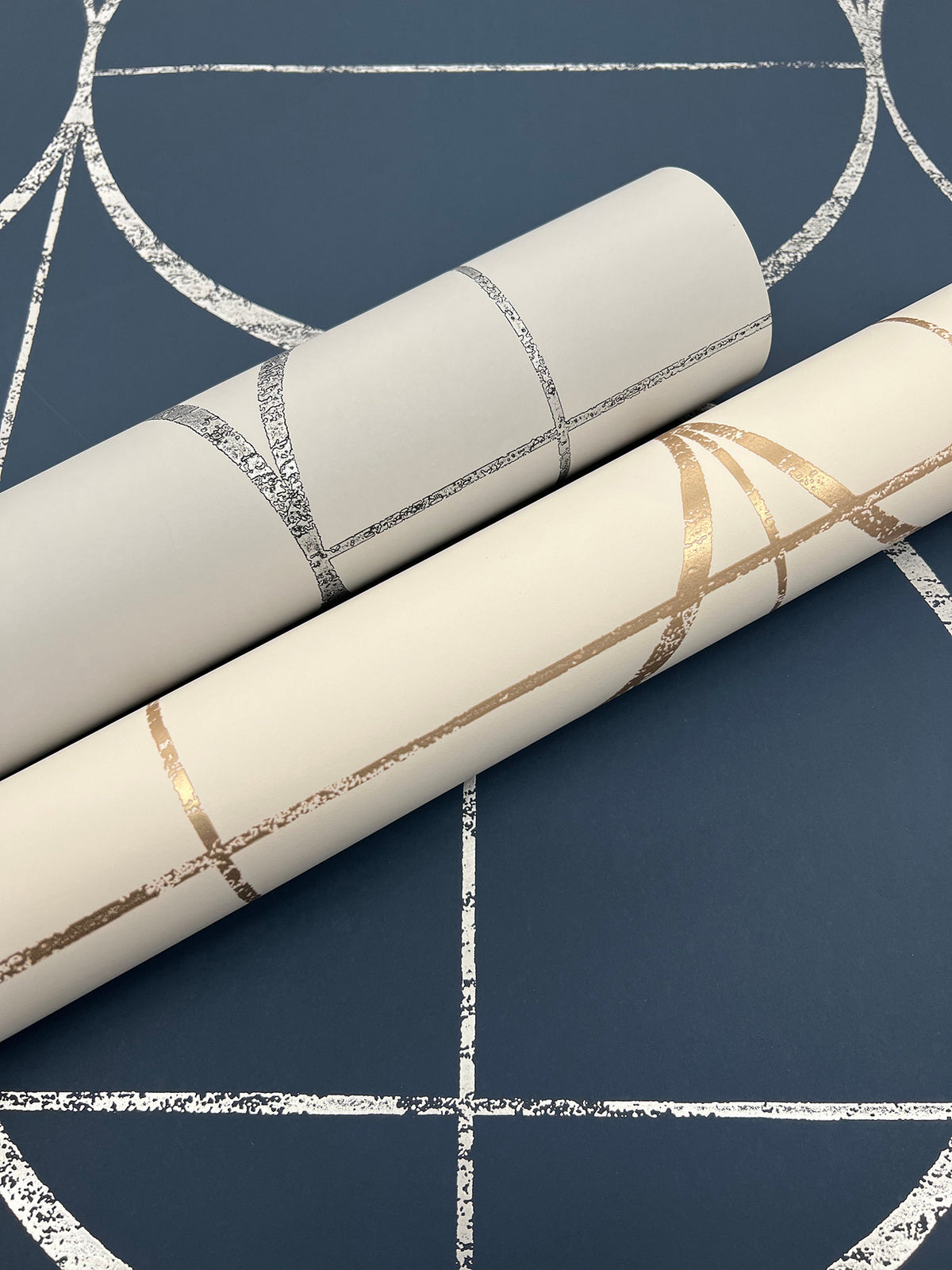 Two rolls of Sun Circles Wallpaper by York Wallcoverings—one in silver and one in gold on cream—rest on a dark blue background with metallic geometric line patterns.