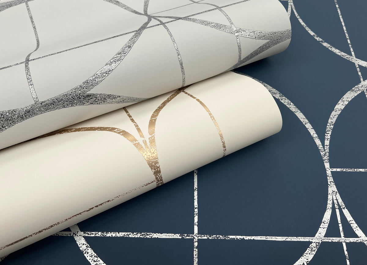 Two rolls of York Wallcoverings’ Sun Circles Wallpaper, featuring metallic geometric line patterns in silver and gold, are placed on top of a dark blue wallpaper displaying a similar silver geometric design.