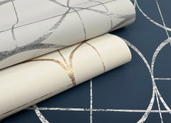 Two rolls of York Wallcoverings’ Sun Circles Wallpaper, featuring metallic geometric line patterns in silver and gold, are placed on top of a dark blue wallpaper displaying a similar silver geometric design.