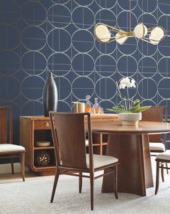 A modern dining room featuring a wooden table, four chairs, a sideboard, contemporary light fixture, a white orchid centerpiece, and York Wallcoverings’ Sun Circles Wallpaper in navy with geometric patterns.