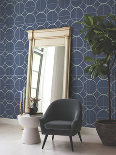 A modern room featuring the Sun Circles Wallpaper by York Wallcoverings in a blue geometric design, a large ornate mirror, a gray armchair, a round white side table adorned with candles and a plant, and a tall potted plant in the corner.