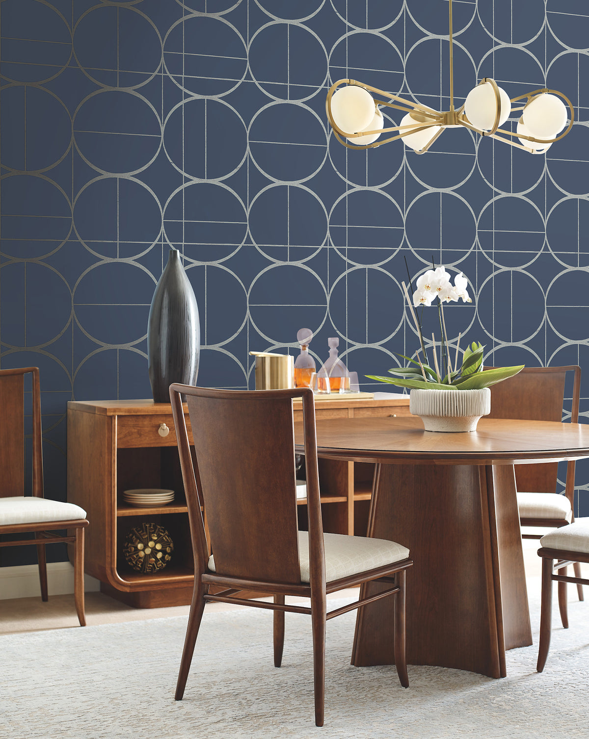 A modern dining room featuring a wooden table and chairs, York Wallcoverings’ Sun Circles Wallpaper in blue with geometric patterns, a gold chandelier, a white orchid centerpiece, and stylish decor on the sideboard.