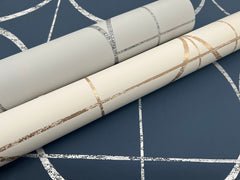 Two rolls of Sun Circles Wallpaper by York Wallcoverings, featuring metallic and textured line designs—one in silver on light gray and the other in gold on cream—are displayed on a blue background with a coordinating white abstract line pattern.