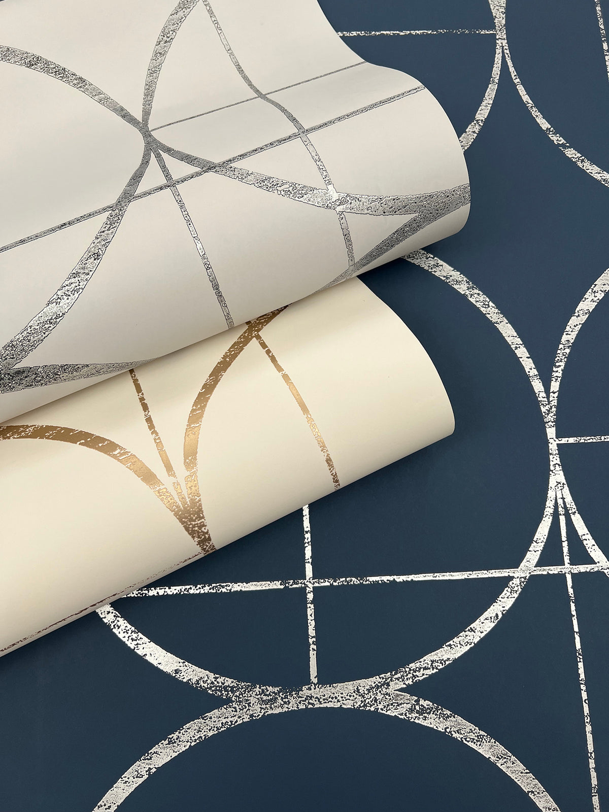 Three rolls of York Wallcoverings Sun Circles Wallpaper feature metallic geometric circle patterns: one roll in navy blue with silver accents, and two rolls in cream with gold and silver designs.