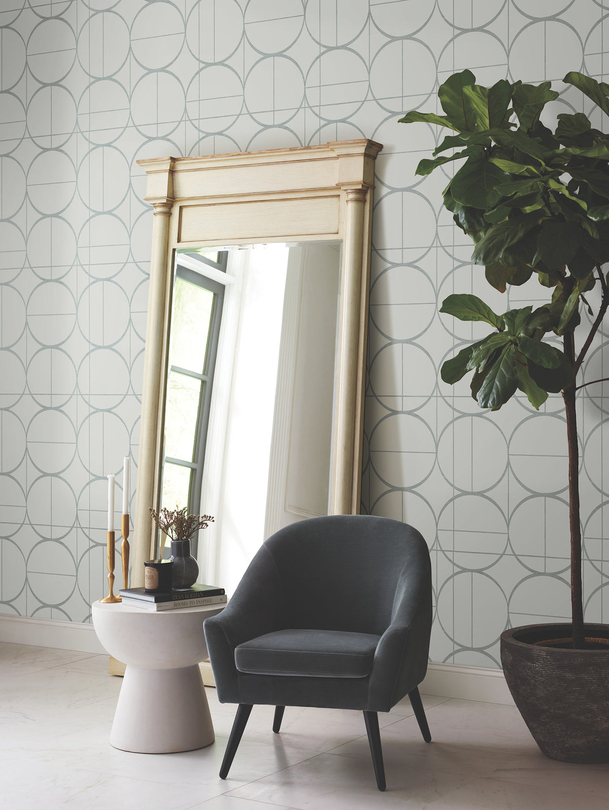 A modern lounge featuring the Sun Circles Wallpaper by York Wallcoverings, a tall mirror with a cream frame, a gray armchair, a small round table with decor, and a large potted plant.