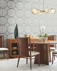 Mid-century modern dining room with wood furniture, a brass chandelier, and a white orchid centerpiece on the table. The walls feature York Wallcoverings’ Sun Circles Wallpaper with geometric patterns, and a sideboard with decor items is in the background.
