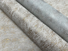 Two rolls of York Wallcoverings’ Shimmering Patina Wallpaper, featuring metallic gold and silver patterns on a light background, are unrolled slightly on a matching surface.