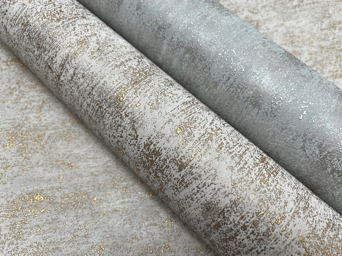 Two rolls of York Wallcoverings’ Shimmering Patina Wallpaper in silver and gold tones lie side by side on a coordinating background, showcasing a subtle metallic sheen and gently distressed pattern.