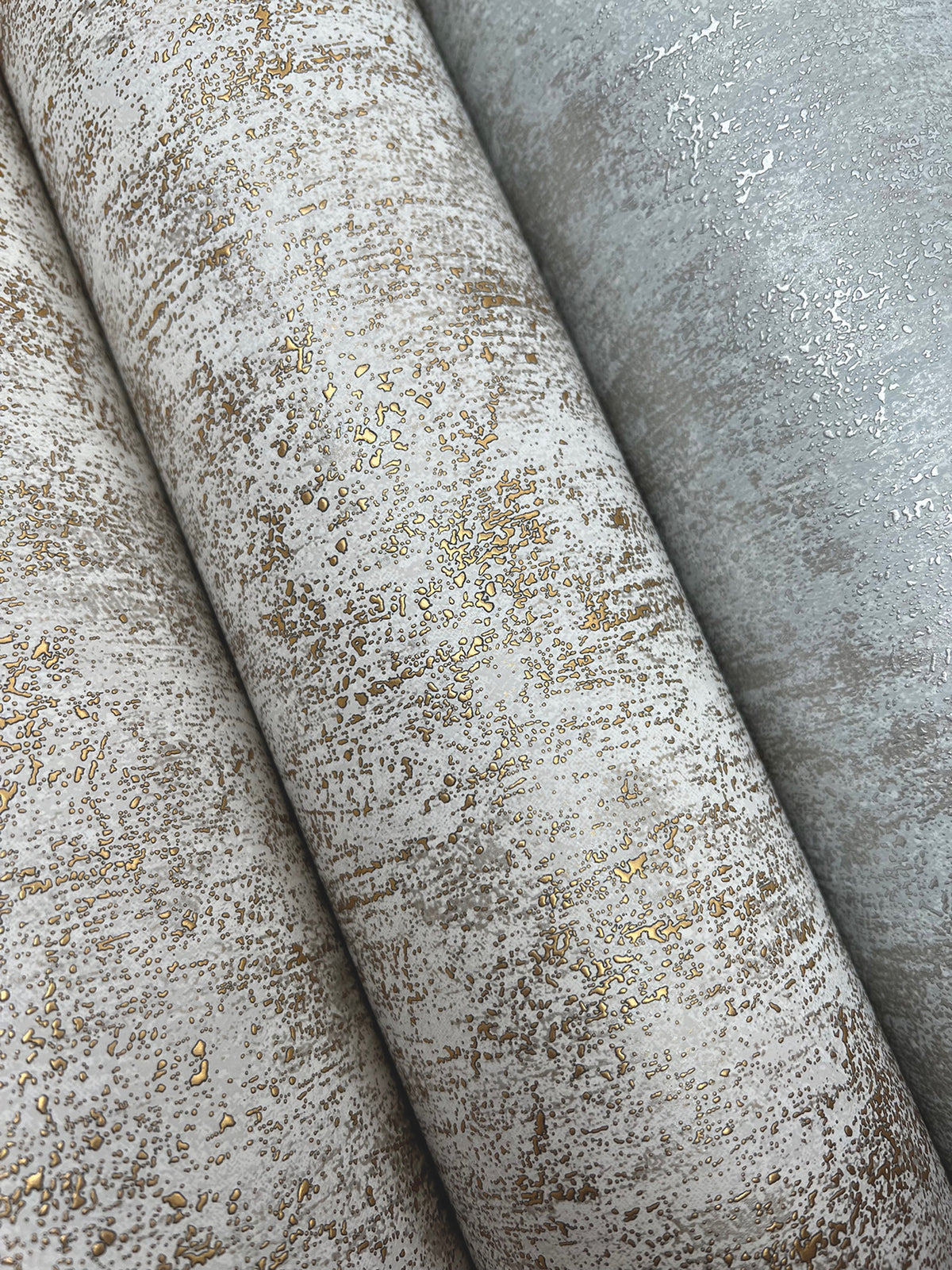 Three rolls of Shimmering Patina Wallpaper by York Wallcoverings, featuring an abstract, distressed pattern in beige and gray tones highlighted with metallic gold accents.