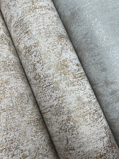 Three rolls of Shimmering Patina Wallpaper by York Wallcoverings, featuring an abstract, distressed pattern in beige and gray tones highlighted with metallic gold accents.