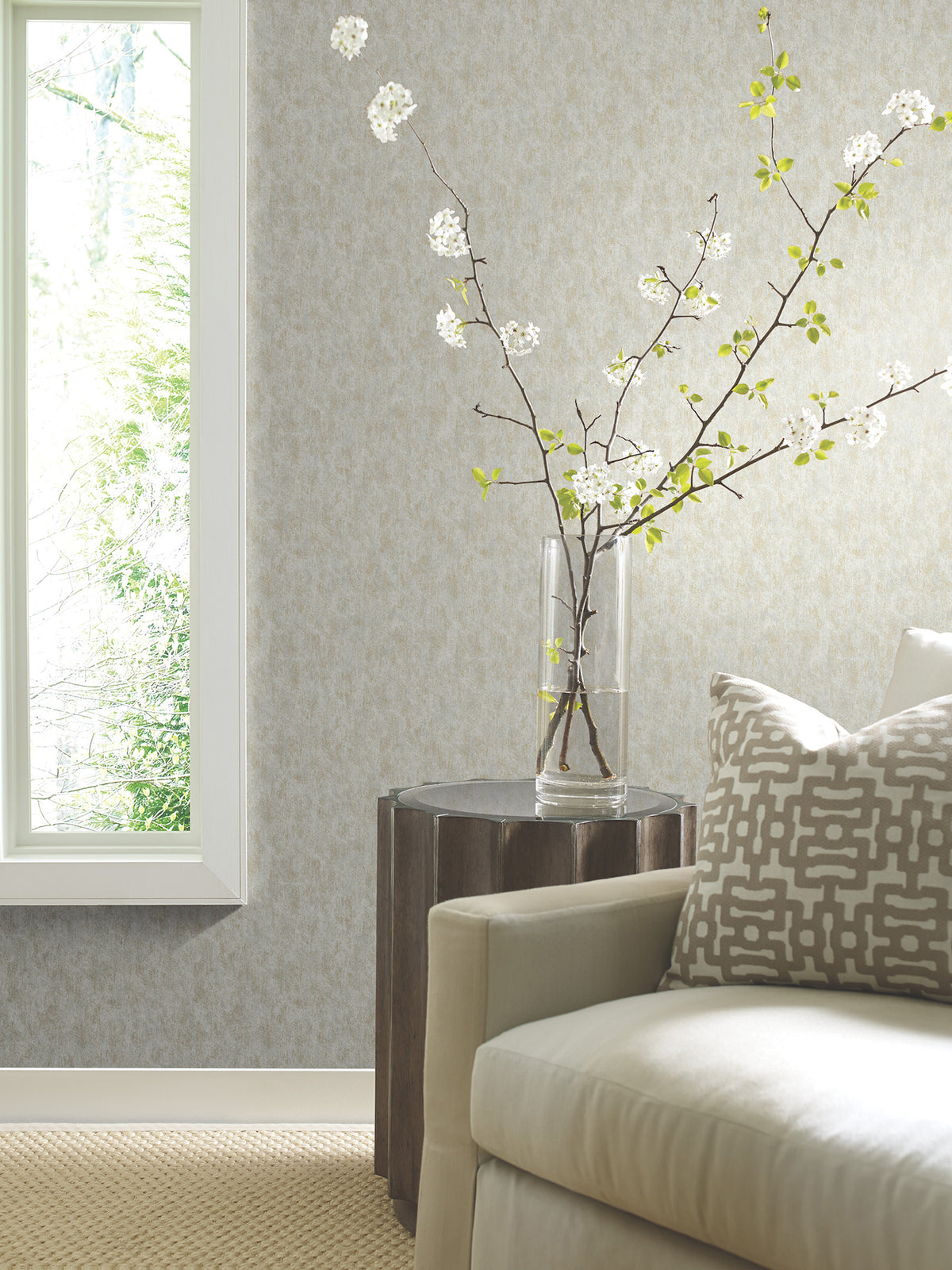 A glass vase with flowering branches sits on a wooden side table next to a light-colored sofa adorned with a patterned pillow. Sunlight streams through a window on the left, illuminating the softly textured Shimmering Patina Wallpaper by York Wallcoverings and the woven carpet.