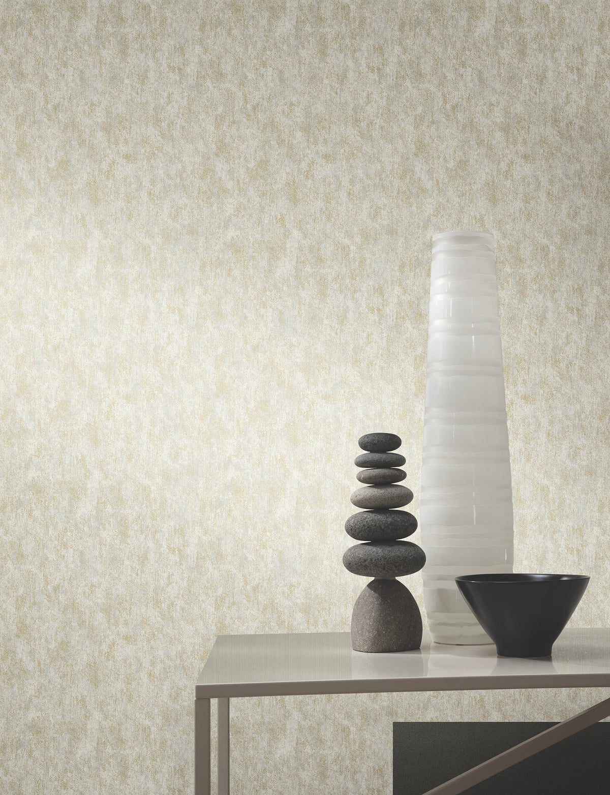 A minimalist table holds a tall white vase, a small black bowl, and stacked smooth stones, set against a beige Shimmering Patina Wallpaper by York Wallcoverings.