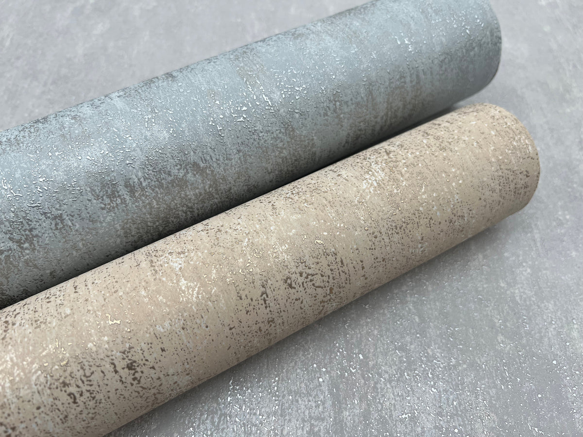 Two rolls of York Wallcoverings’ Shimmering Patina Wallpaper, one in light blue and the other in beige, lie side by side on a gray surface. Each roll features a subtle, metallic speckled pattern.
