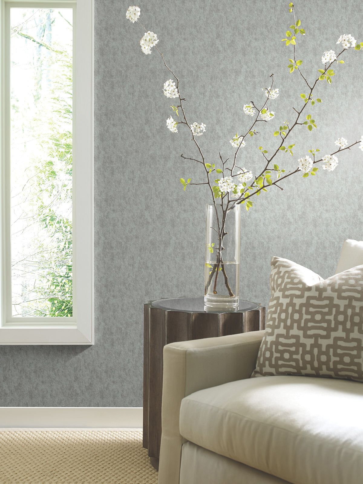 A modern living room featuring York Wallcoverings’ Shimmering Patina Wallpaper, complemented by a light sofa, geometric pillow, wooden side table, glass vase with flowering branches, beige carpet, and a large window filling the space with natural light.