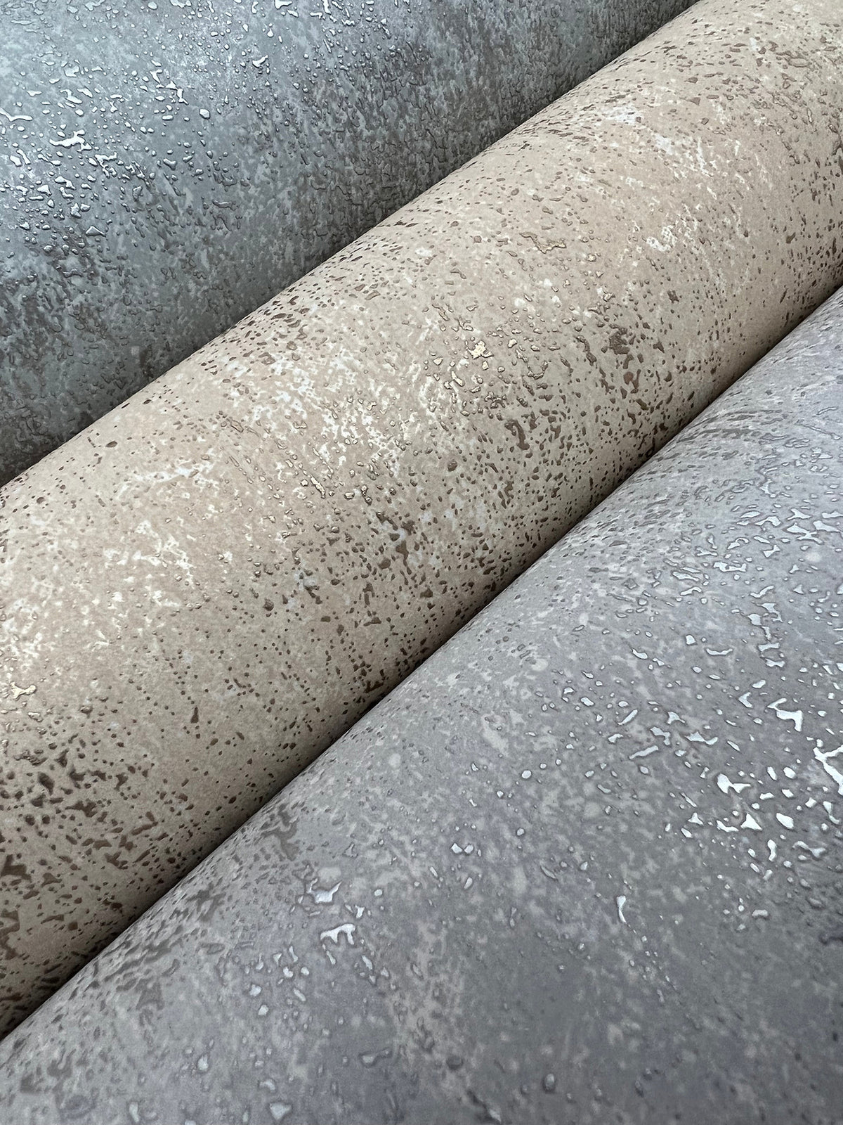 Three rolls of Shimmering Patina Wallpaper by York Wallcoverings in gray, beige, and taupe shades are shown up close. The wallpaper features a slightly reflective, irregular pattern reminiscent of water droplets or weathered surfaces.