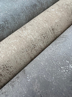 Three rolls of Shimmering Patina Wallpaper by York Wallcoverings in gray, beige, and taupe shades are shown up close. The wallpaper features a slightly reflective, irregular pattern reminiscent of water droplets or weathered surfaces.