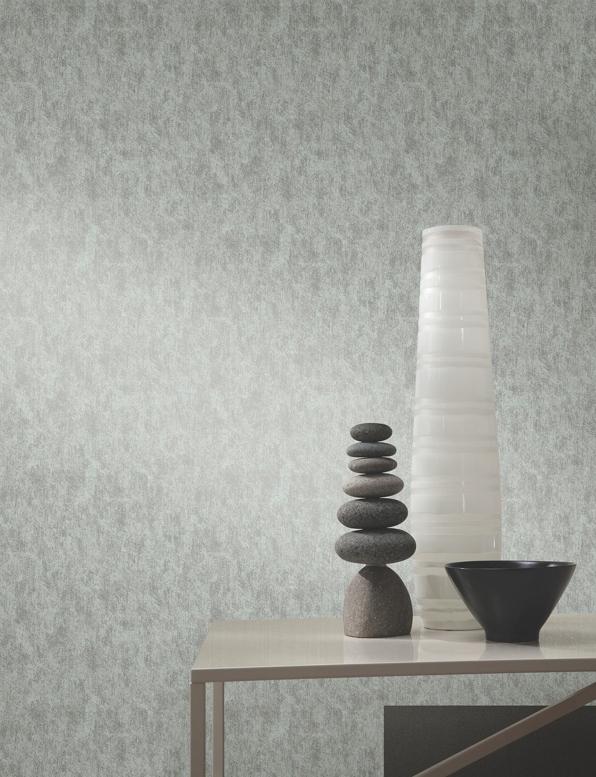 A modern table with a black bowl, stacked smooth stones, and a tall white vase sits against a wall adorned with York Wallcoverings’ Shimmering Patina Wallpaper.