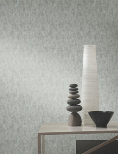 A modern table with a black bowl, stacked smooth stones, and a tall white vase sits against a wall adorned with York Wallcoverings’ Shimmering Patina Wallpaper.