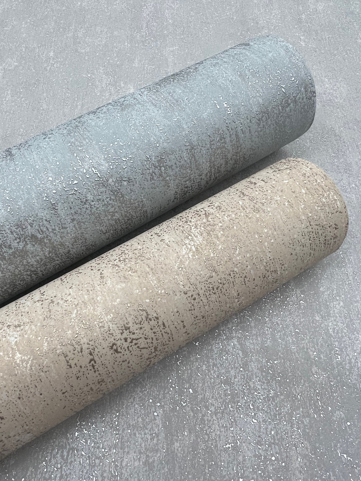 Two rolls of York Wallcoverings’ Shimmering Patina Wallpaper—one in light grey and one in beige—are positioned parallel to each other on a sparkly, light-colored surface.