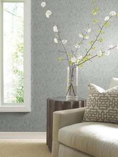 A modern living room corner featuring a beige sofa, a patterned pillow, a round wooden side table, and a clear vase with flowering branches; the walls are adorned with York Wallcoverings’ Shimmering Patina Wallpaper in light gray, illuminated by natural light from the window.