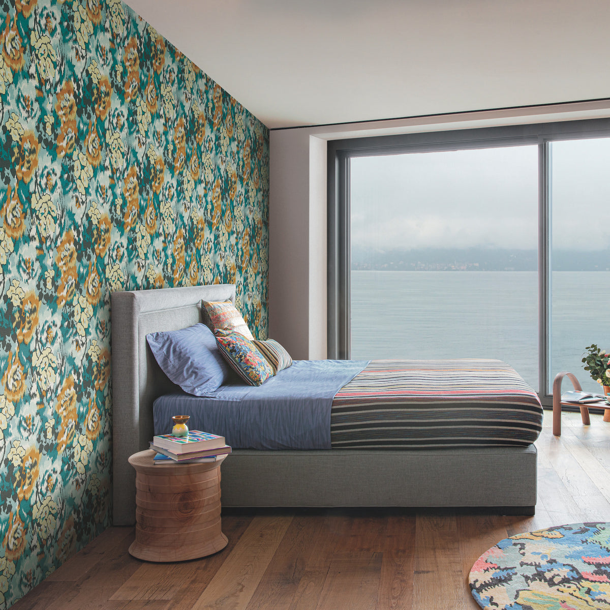 Modern bedroom featuring York Wallcoverings’ Flower Pot Wallpaper as a colorful floral accent wall, a bed with blue and striped bedding, a small round wooden side table, and a large window overlooking a body of water beneath a cloudy sky.