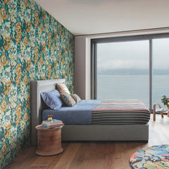 Modern bedroom featuring York Wallcoverings’ Flower Pot Wallpaper as a colorful floral accent wall, a bed with blue and striped bedding, a small round wooden side table, and a large window overlooking a body of water beneath a cloudy sky.