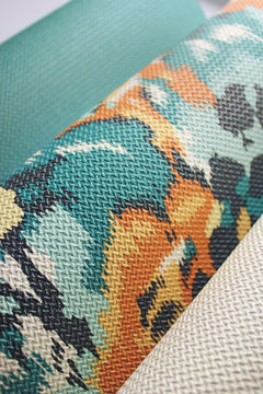 Three textured rolls of York Wallcoverings’ Flower Pot Wallpaper are shown: one in teal, one featuring a teal, orange, black, and yellow abstract pattern, and one in off-white. The wallpapers display a woven texture and are positioned diagonally across the image.