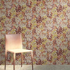 A light pink chair sits on a beige carpet in front of York Wallcoverings’ Flower Pot Wallpaper, which features a floral pattern in shades of pink, brown, and yellow.