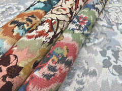 Three rolls of Flower Pot Wallpaper by York Wallcoverings, featuring colorful floral patterns, rest on a surface covered with the same wallpaper design in muted, pale tones. The distinctive textures and vibrant motifs are clearly showcased.