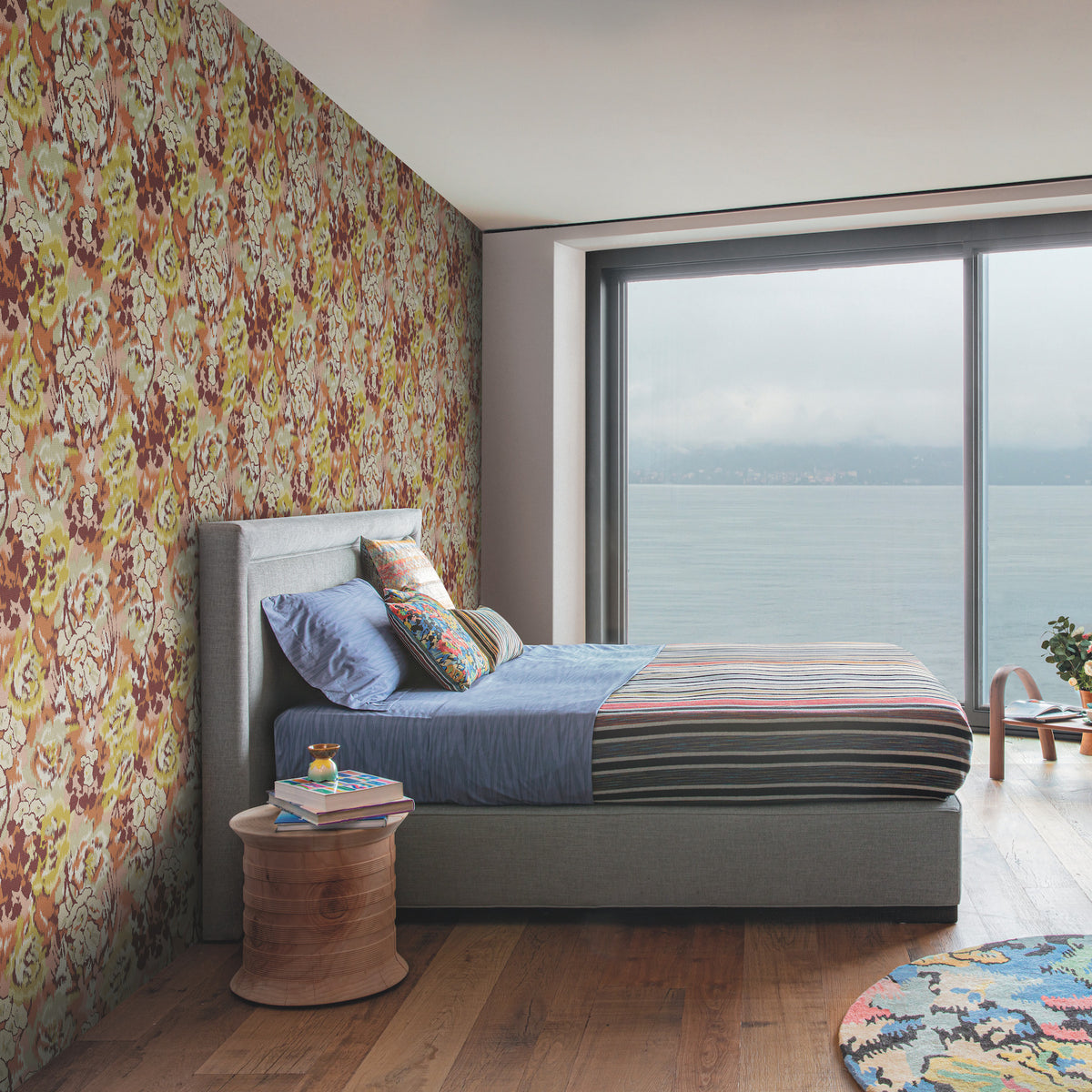 A modern bedroom featuring York Wallcoverings’ Flower Pot Wallpaper as a floral accent wall, with a gray bed frame, blue bedding, striped blanket, round wooden side table, large window, and a scenic water view.