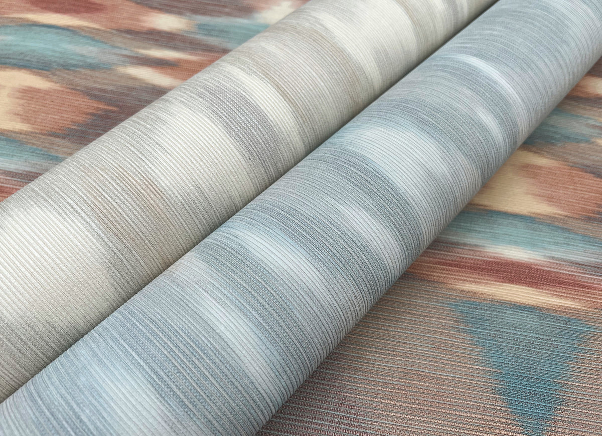 Two rolls of Sunrise Flame Wallpaper by York Wallcoverings, featuring muted blue and beige tones with subtle striped patterns, lying side by side on a similarly colored and patterned surface.