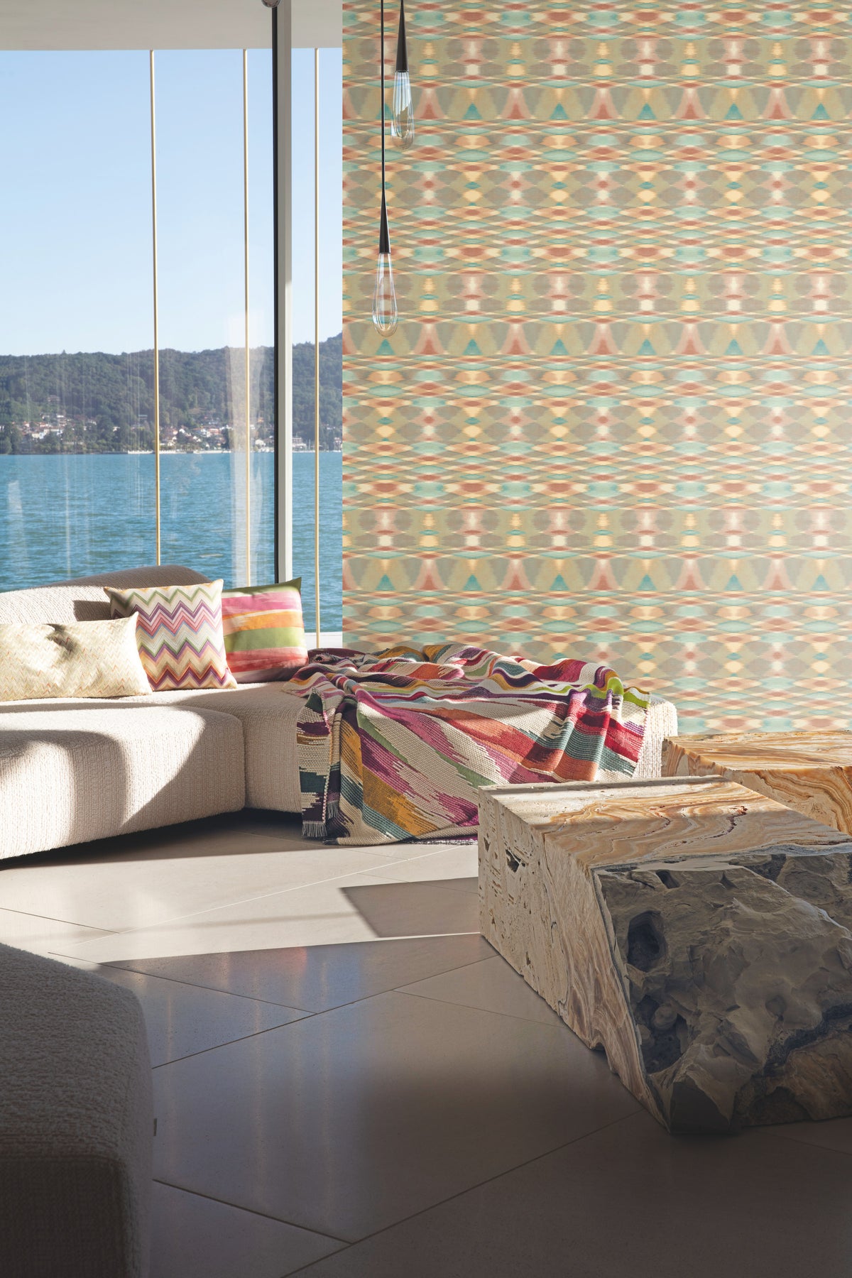A sunlit living room with large windows features a white sectional sofa adorned with colorful pillows and a throw, a stone coffee table, and a patterned feature wall covered in York Wallcoverings’ Sunrise Flame Wallpaper, overlooking a body of water and forested hills.