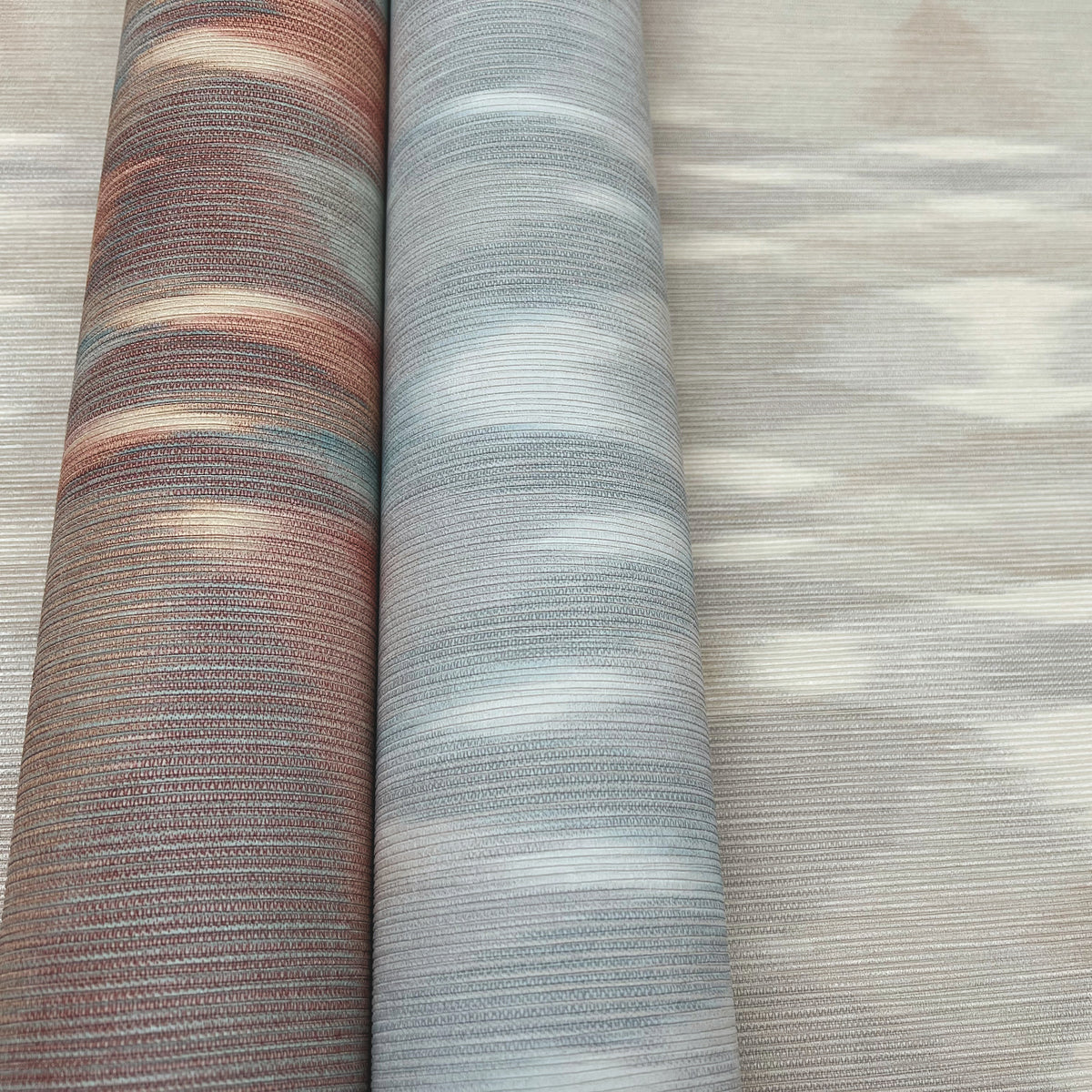 Two rolled-up bolts of York Wallcoverings’ Sunrise Flame Wallpaper—one in earthy brown and the other in pale blue, both featuring subtle horizontal stripes—are displayed side by side on a neutral-patterned surface.