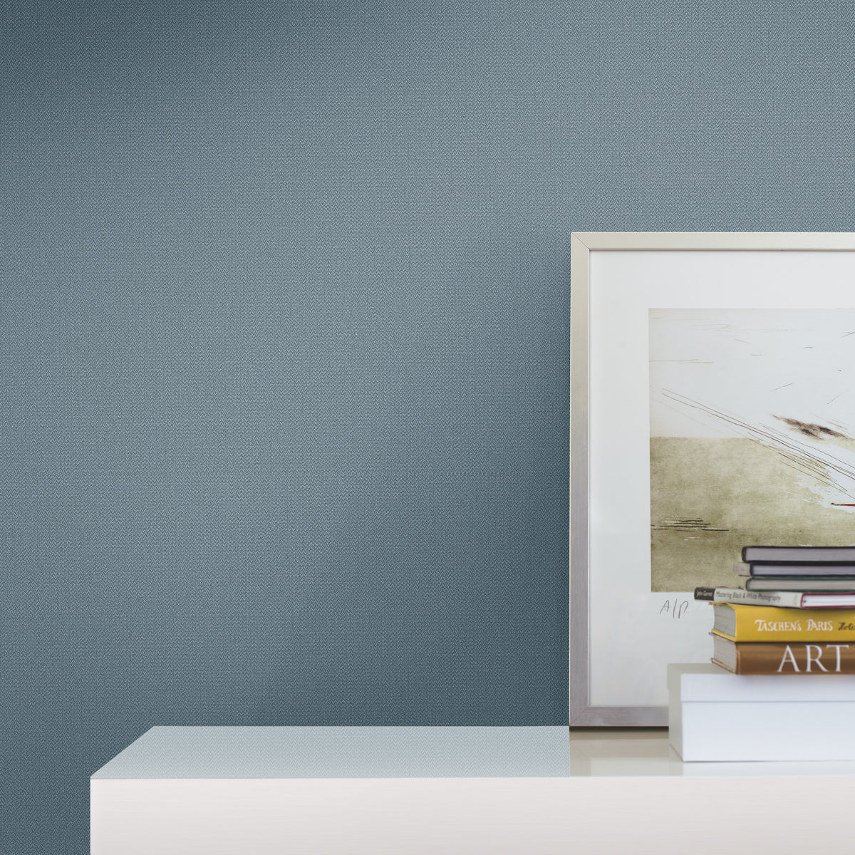 On a white shelf set against Chevronette Wallpaper by York Wallcoverings in a light blue hue, a framed abstract artwork, stacked books, and a closed notebook are displayed.