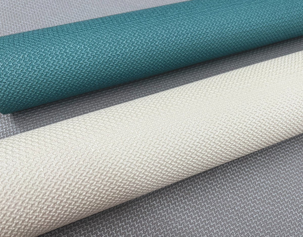 Three rolls of Chevronette Wallpaper by York Wallcoverings are displayed at an angle, featuring teal, cream, and light gray colors. The wallpapers have a woven texture and are laid on a gray, textured surface.