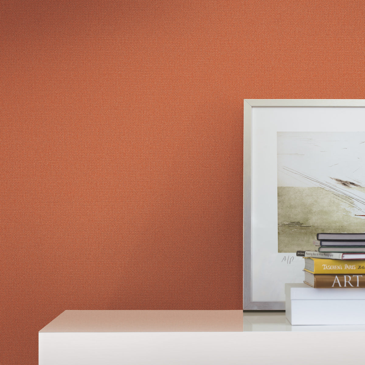 A framed abstract art print, stacked books, and a white box sit on a glossy white surface against a textured burnt orange wall covered in York Wallcoverings’ Chevronette Wallpaper.