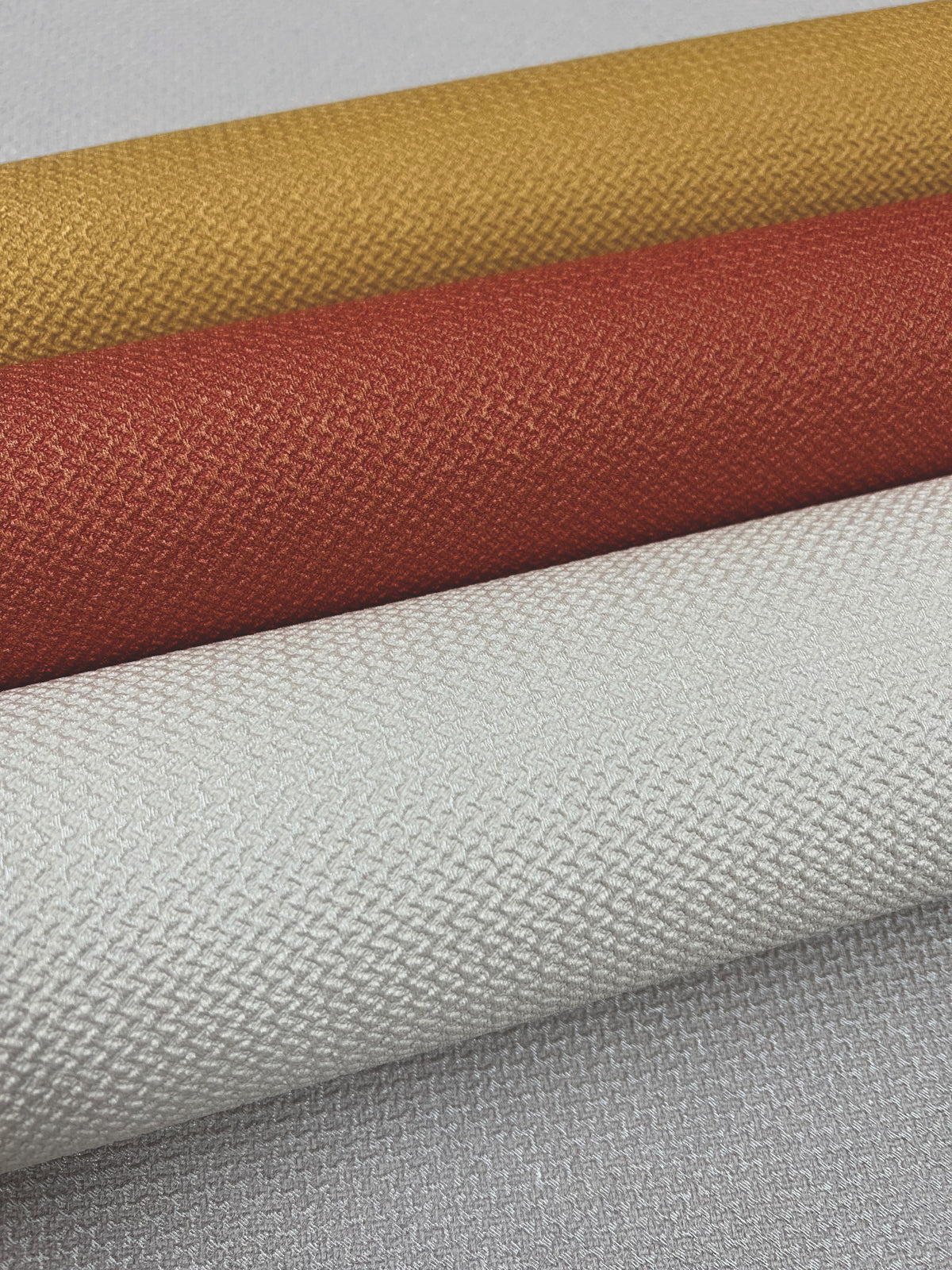 Four rolls of York Wallcoverings’ Chevronette Wallpaper in mustard yellow, burnt orange, and two shades of light gray are stacked diagonally, displaying their woven-inspired patterns.