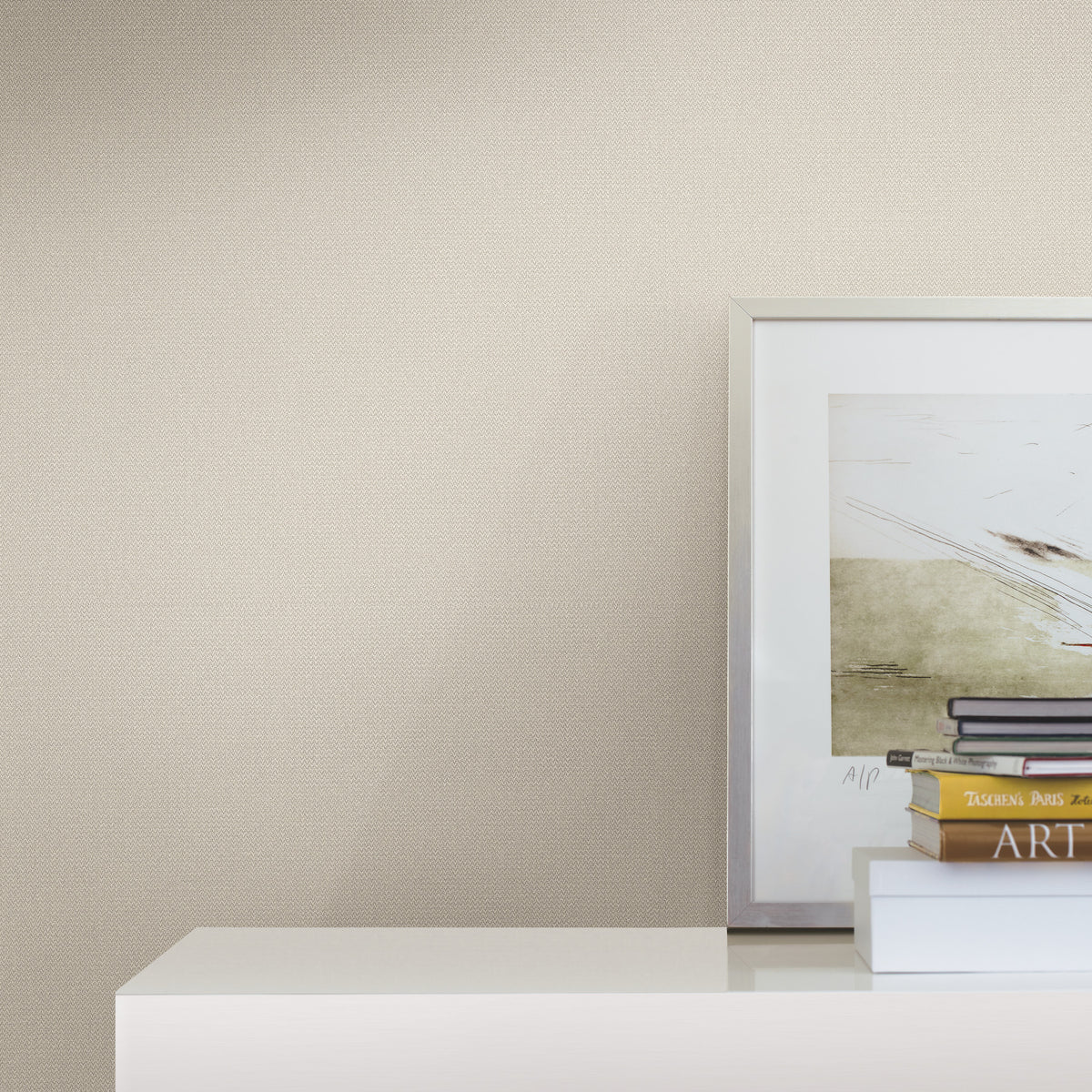 A framed abstract painting and a stack of books sit on a white shelf against a beige Chevronette Wallpaper by York Wallcoverings.
