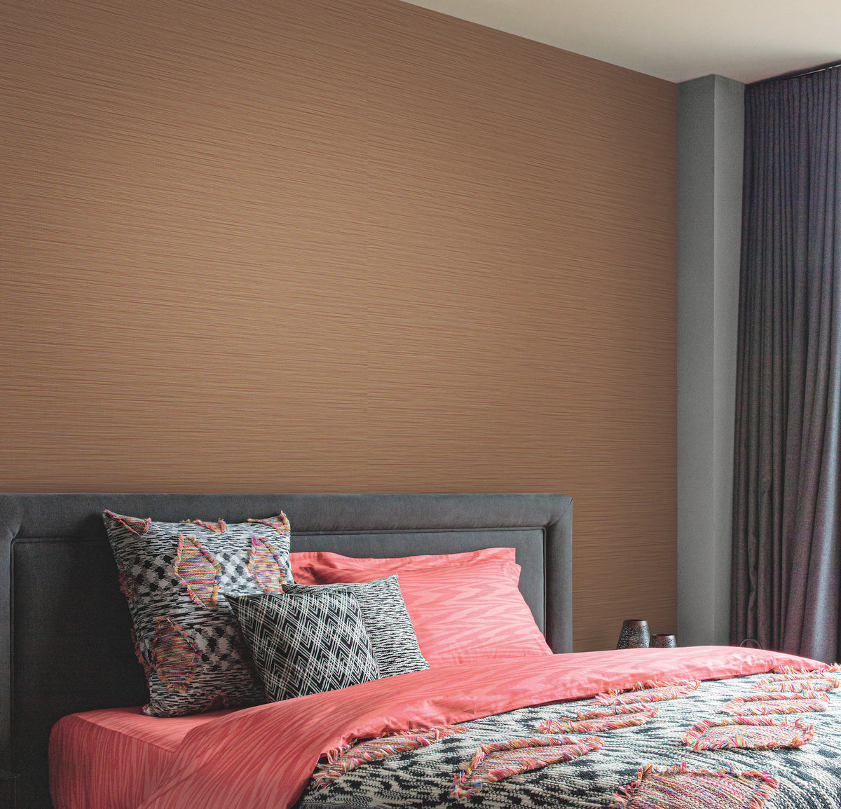 A modern bedroom featuring a padded dark gray headboard, coral pink bedding with abstract patterns, patterned pillows, and the Cannete Wallpaper by York Wallcoverings in light brown, complemented by dark floor-length curtains.