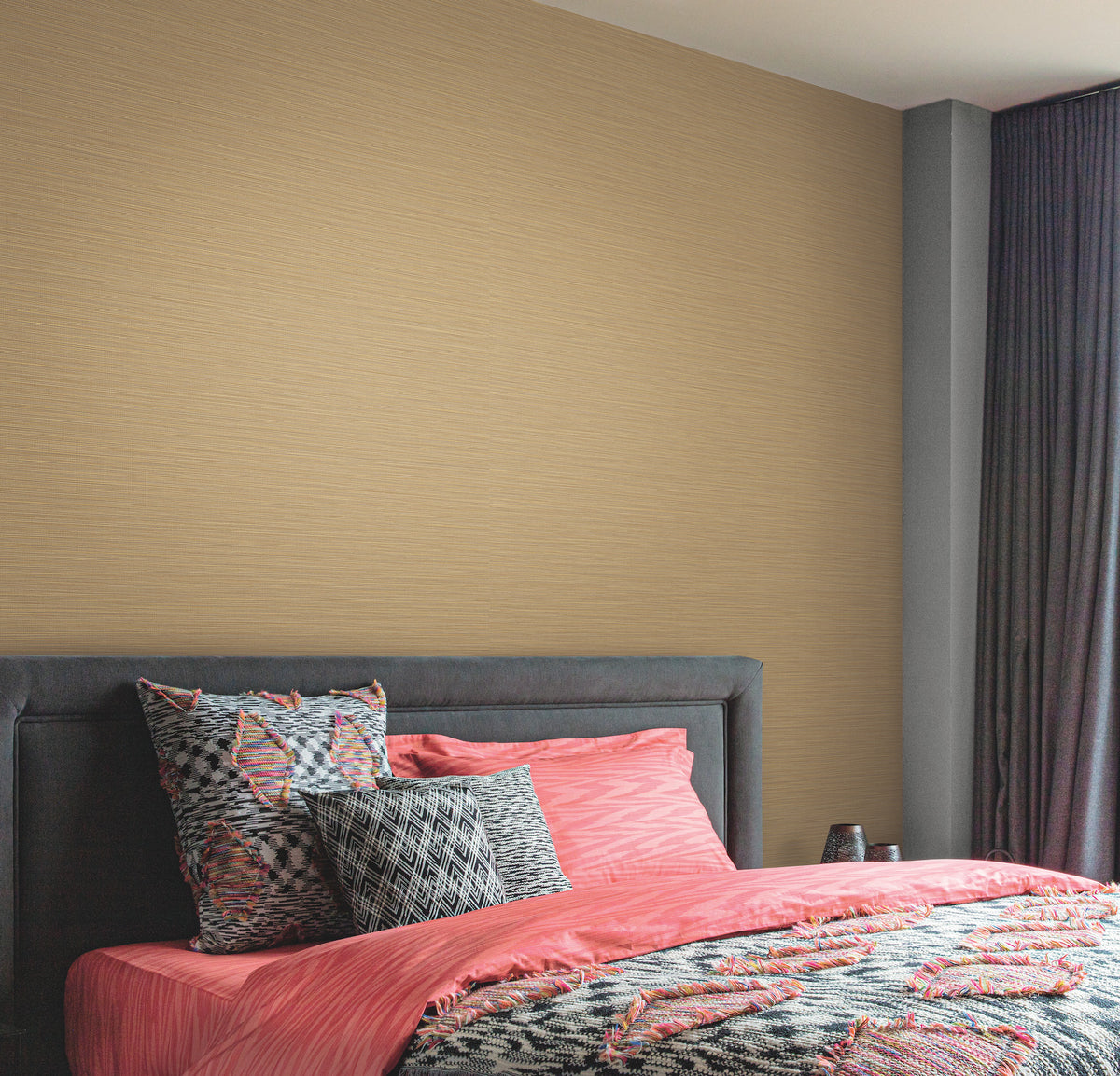 A modern bedroom featuring the Cannete Wallpaper by York Wallcoverings in a neutral beige textured finish, with a gray upholstered headboard, coral and gray geometric bedding, and dark curtains framing the window on the right.